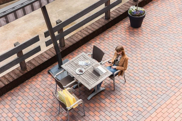 Vista aérea de la mujer con teléfono inteligente cerca de la computadora portátil, café y compras en la terraza - foto de stock
