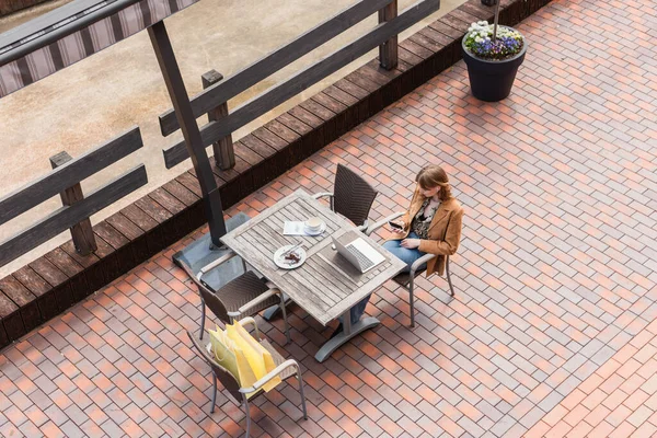 Overhead view of woman using smartphone near laptop, coffee and purchases on terrace — Stock Photo