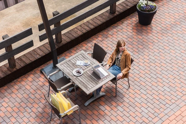 Overhead view of stylish woman sitting near gadgets, dessert and shopping bags on terrace — Stock Photo