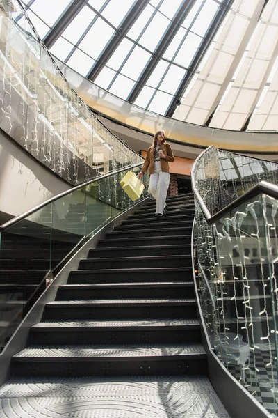 Низкий угол зрения улыбающийся клиент со смартфоном и сумки ходить по лестнице в магазине — стоковое фото