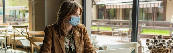 Стильная женщина в медицинской маске сидит рядом с ноутбуком в кафе, баннер — стоковое фото