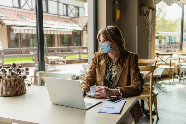Mujer con máscara médica sentada cerca de aparatos y menú en la cafetería - foto de stock