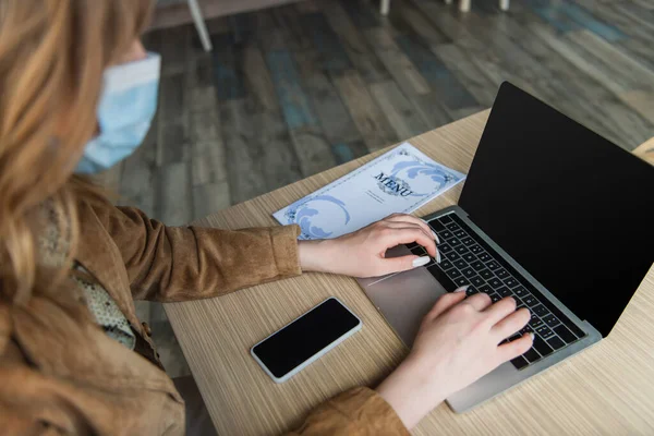 Blurred woman in medical mask using laptop near cellphone with blank screen and menu in cafe — Stock Photo