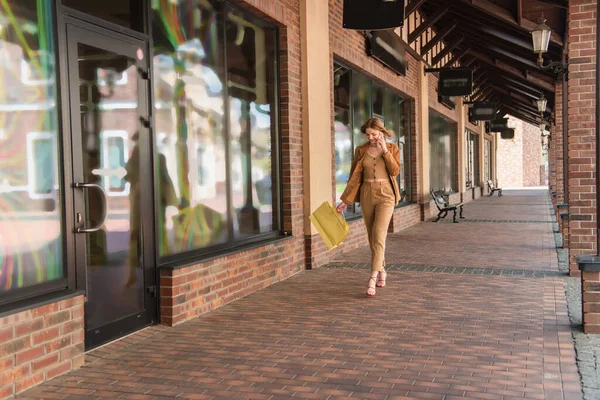 Lächelnde Frau mit Einkaufstüten spricht auf Smartphone in der Nähe des Gebäudes — Stockfoto