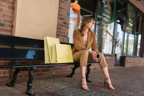 Вид сбоку стильной женщины со смартфоном, сидящей на скамейке рядом с сумками для покупок — стоковое фото