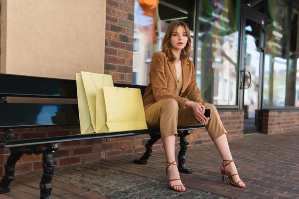 Hübsche Frau hält Handy neben Gebäude und Einkaufstüten auf Bank im Freien — Stockfoto