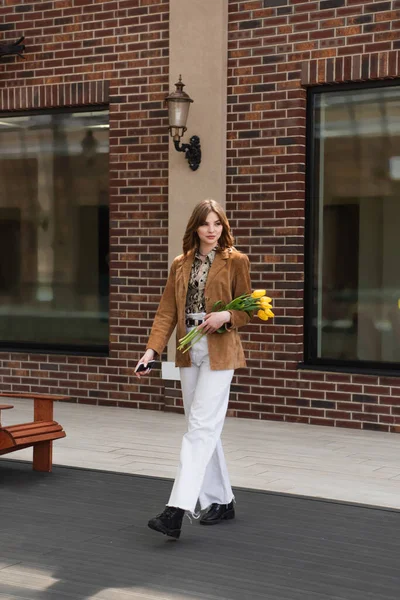 Femme élégante avec smartphone tenant des tulipes et marchant à l'extérieur — Photo de stock