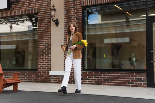 Mulher elegante com copo de papel segurando tulipas e andando ao ar livre — Fotografia de Stock