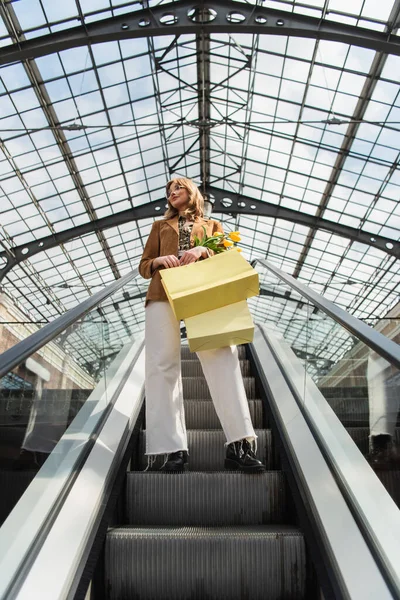 Full length of woman in sunglasses holding yellow tulips and shopping bags while standing on escalator — Stock Photo