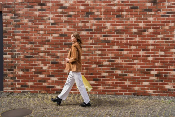 Longitud completa de la joven mujer con estilo en la chaqueta caminando con bolsas de compras cerca de la pared de ladrillo - foto de stock