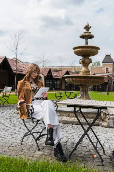 Стильная женщина в солнечных очках с помощью цифрового планшета и с бумажной чашкой в руках, сидя в открытом кафе — стоковое фото