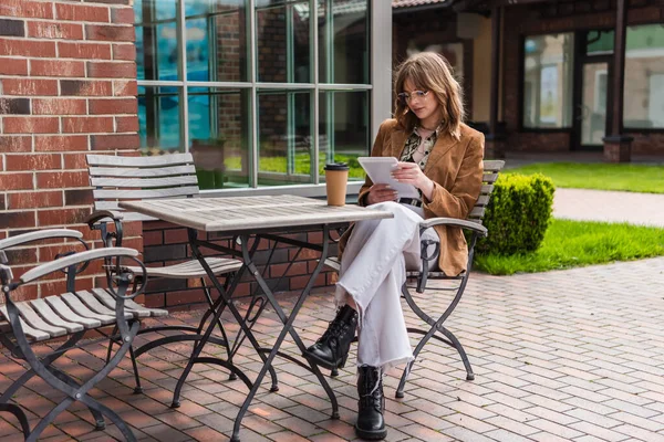 Comprimento total da mulher elegante em óculos de sol e jaqueta segurando tablet digital perto de copo de papel na mesa — Fotografia de Stock