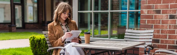 Femme élégante dans des lunettes de soleil et veste tenant tablette numérique près de tasse en papier sur la table, bannière — Photo de stock