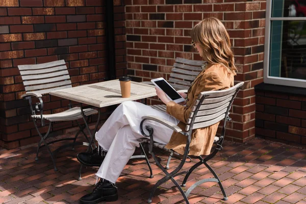 Longitud completa de la mujer de moda en gafas de sol y chaqueta que sostiene la tableta digital con pantalla en blanco cerca de la taza de papel en la mesa - foto de stock