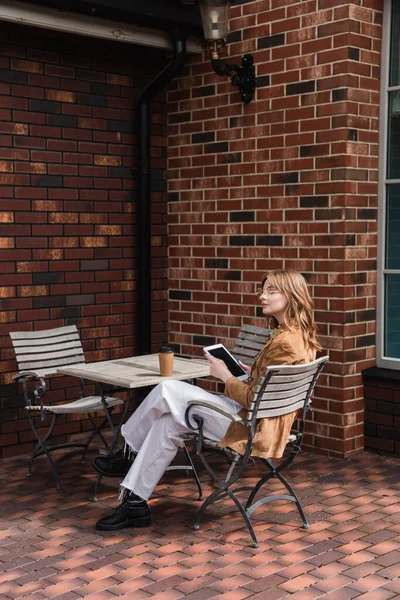 Pleine longueur de femme élégante dans des lunettes de soleil et veste tenant tablette numérique avec écran blanc près de tasse de papier sur la table — Photo de stock