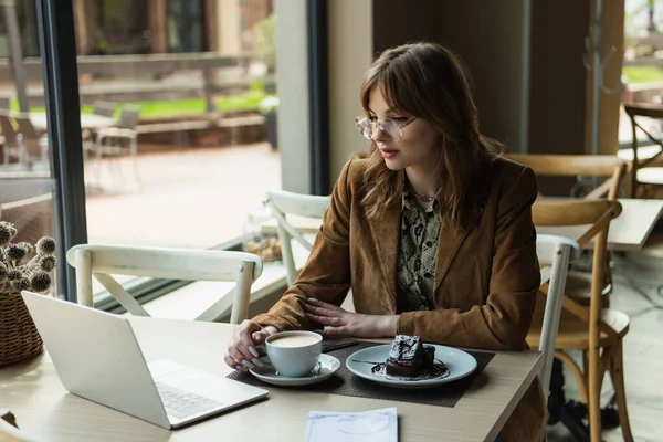 Молодая женщина в очках смотрит на ноутбук возле торта и чашку капучино в кафе — стоковое фото
