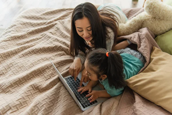 Vista aérea de mujer asiática alegre usando el ordenador portátil cerca de la hija en la cama - foto de stock