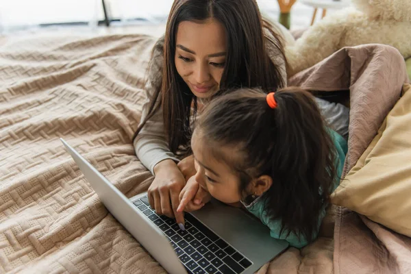 Asiatisches Mädchen zeigt auf Laptop in der Nähe von Mutter auf Bett — Stockfoto
