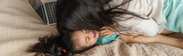 Мати цілує азіатську дитину біля ноутбука на ліжку, банер — стокове фото