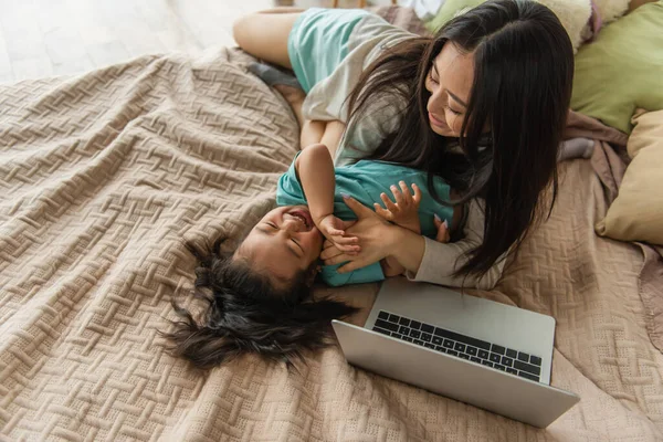 Vista aerea della madre che si diverte con il bambino asiatico vicino al computer portatile sul letto — Foto stock