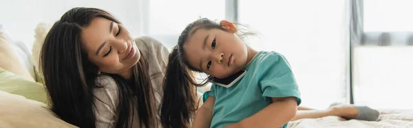 Lächelnde Frau schaut asiatische Tochter an, die im Schlafzimmer auf dem Handy spricht, Banner — Stockfoto