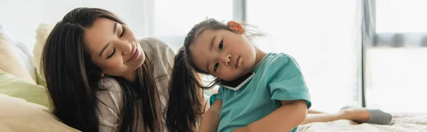 Donna sorridente guardando la figlia asiatica che parla sul cellulare in camera da letto, banner — Foto stock