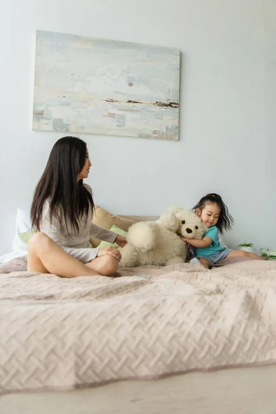 Mãe asiática sentada perto da filha com brinquedo macio na cama — Fotografia de Stock