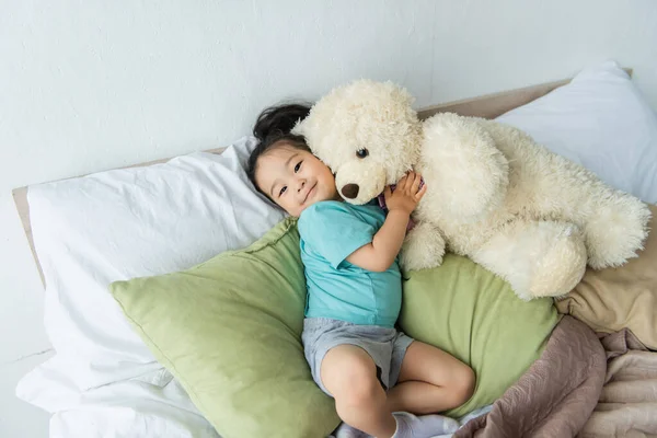 Smiling asian kid holding teddy bear on bed — Stock Photo