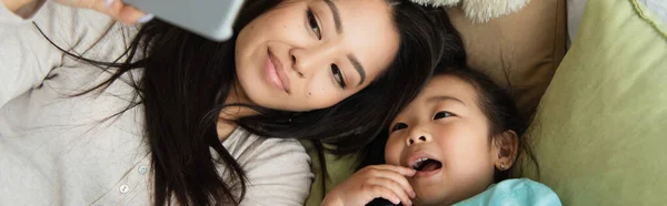 Alto ángulo vista de asiático madre e hija tomando selfie en cama, bandera - foto de stock