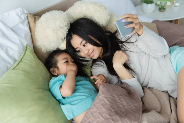 Sonriente madre con smartphone mirando asiático niño en la cama - foto de stock