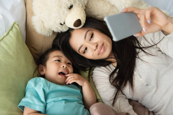 High angle view of mother and asian kid taking selfie on smartphone near teddy bear on bed — Stock Photo