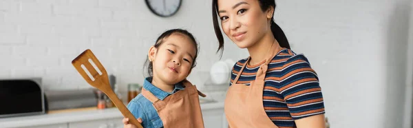 Asiatisches Kind mit Spachtel blickt in die Kamera nahe der Mutter in der Küche, Banner — Stockfoto