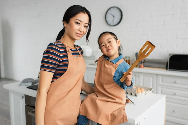 Mujer asiática mirando a la cámara cerca de hija con espátula en la cocina - foto de stock