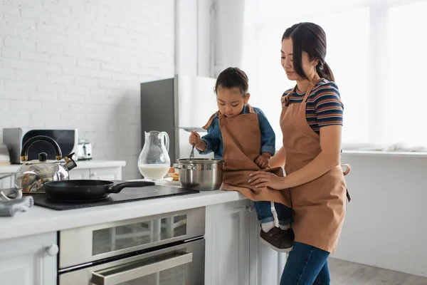 Sonriendo mujer asiática de pie cerca de hija con batidor en la cocina - foto de stock