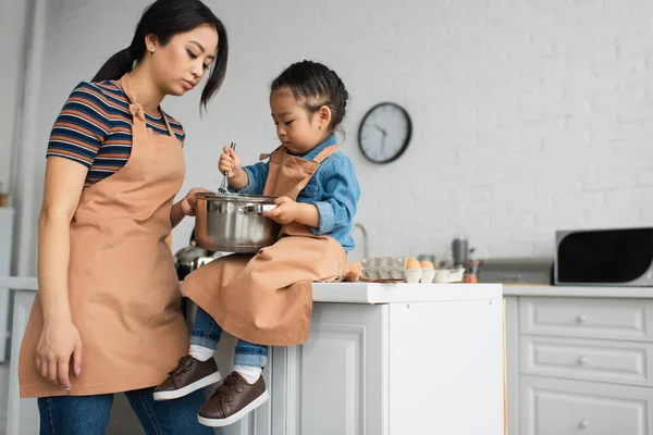 Asiatisches Mädchen hält Pfanne und Schneebesen in der Küche nahe der Mutter — Stockfoto