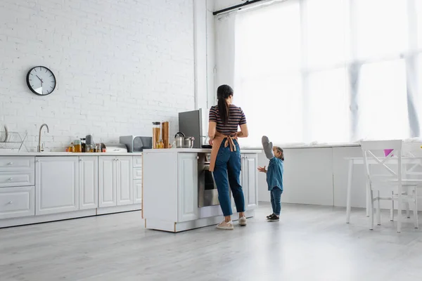 Niño asiático con guante de horno de pie cerca de la madre en la cocina - foto de stock