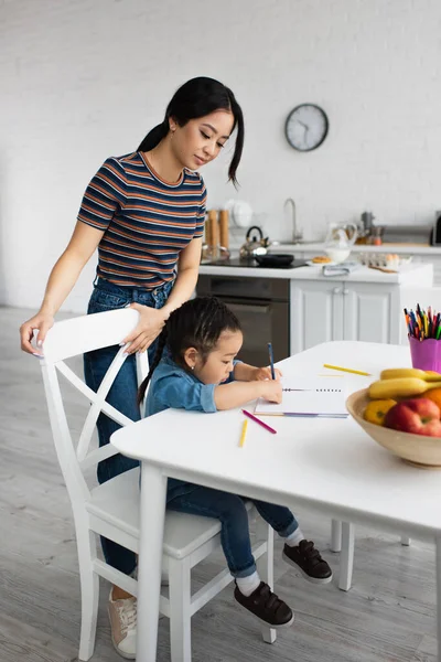 Mãe asiática em pé perto de criança desenho e frutas na mesa — Fotografia de Stock