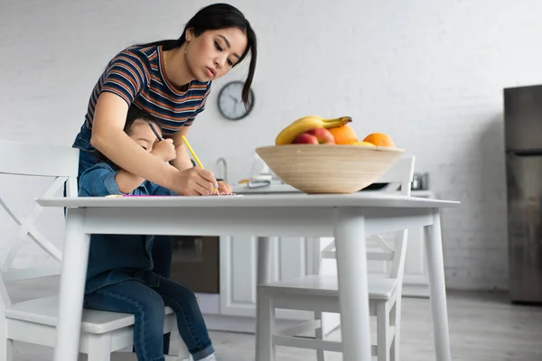 Asian woman drawing near daughter and blurred fruits — Stock Photo