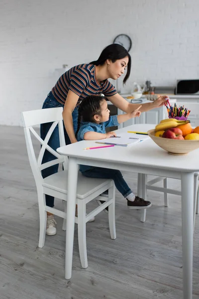 Vista lateral del niño asiático apuntando a lápices de color cerca de la madre y las frutas - foto de stock