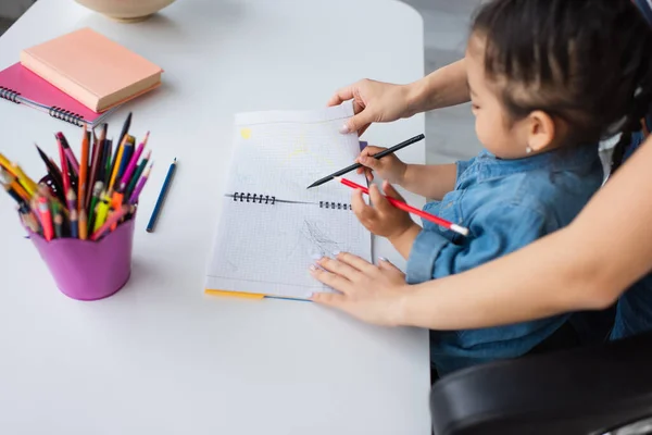 Вид сбоку матери, стоящей рядом с азиатским ребенком с блокнотом и размытыми цветными карандашами — стоковое фото