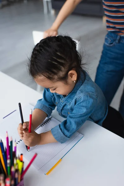 Азійська дівчинка малює близько розмитих кольорових олівців і батьків удома. — стокове фото