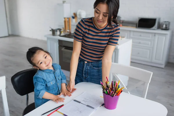 Asiatique fille souriant à la caméra près de notebook, crayons de couleur et mère — Photo de stock