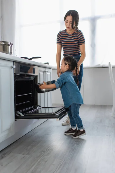 Asiatique enfant mettre casserole dans four près de mère dans la cuisine — Photo de stock