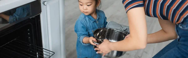 Mother holding pan near asian daughter and oven at home, banner — Stock Photo