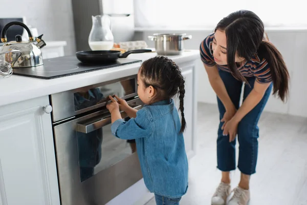 Asiática niño tocando pantalla de horno cerca de madre en cocina - foto de stock