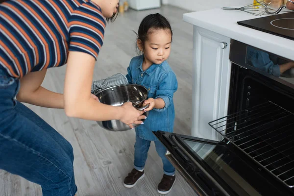 Asiatisches Kind im Ofenhandschuh hält Pfanne in der Nähe von Mutter und Ofen — Stockfoto