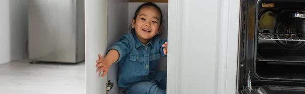 Cheerful asian kid sitting in kitchen cabinet, banner — Stock Photo