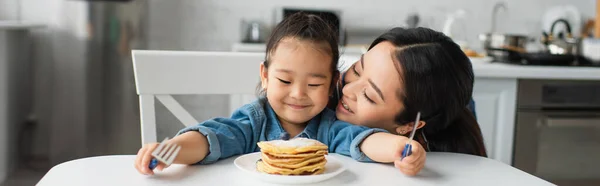 Asian mother kissing child with cutlery near pancakes on table, banner — Stock Photo