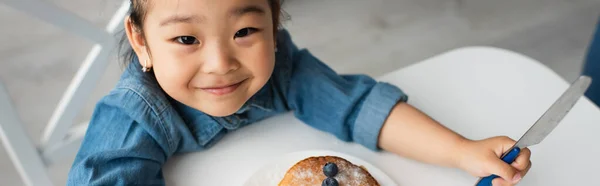 Smiling asian girl with knife looking at camera near pancakes, banner — Stock Photo