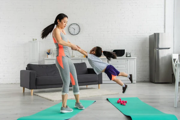 Mulher asiática girando filha perto de tapetes de fitness em casa — Fotografia de Stock